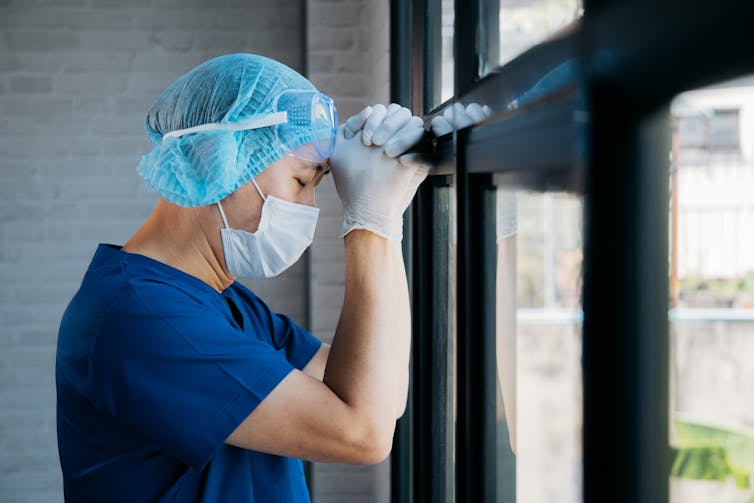 A doctor wearing PPE leaning against a window, appears stressed.